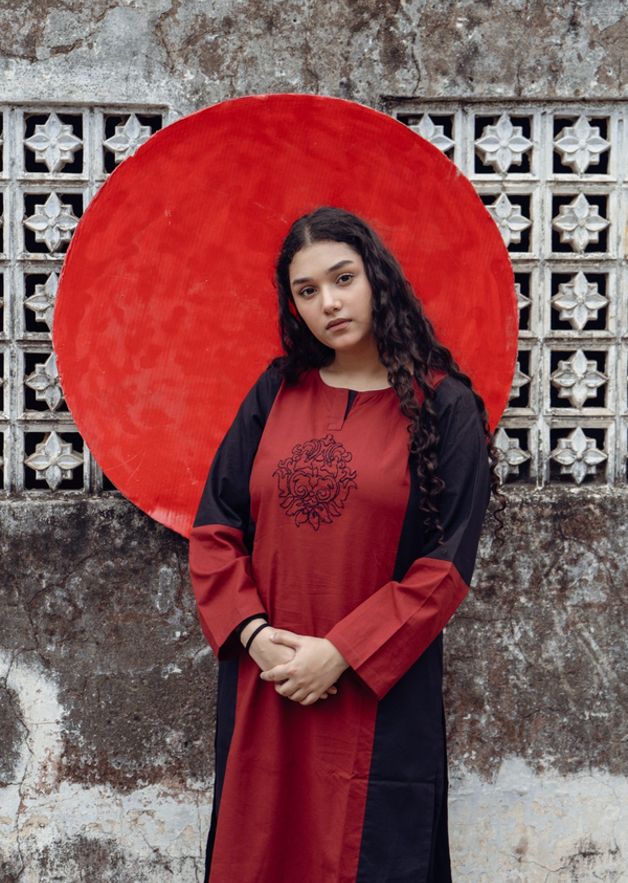 A young woman with long dark curly hair stands in front of a textured wall with a large red circular backdrop, wearing a long-sleeved tunic dress with a color-blocked design in red and black, featuring a black floral embroidery on the chest.